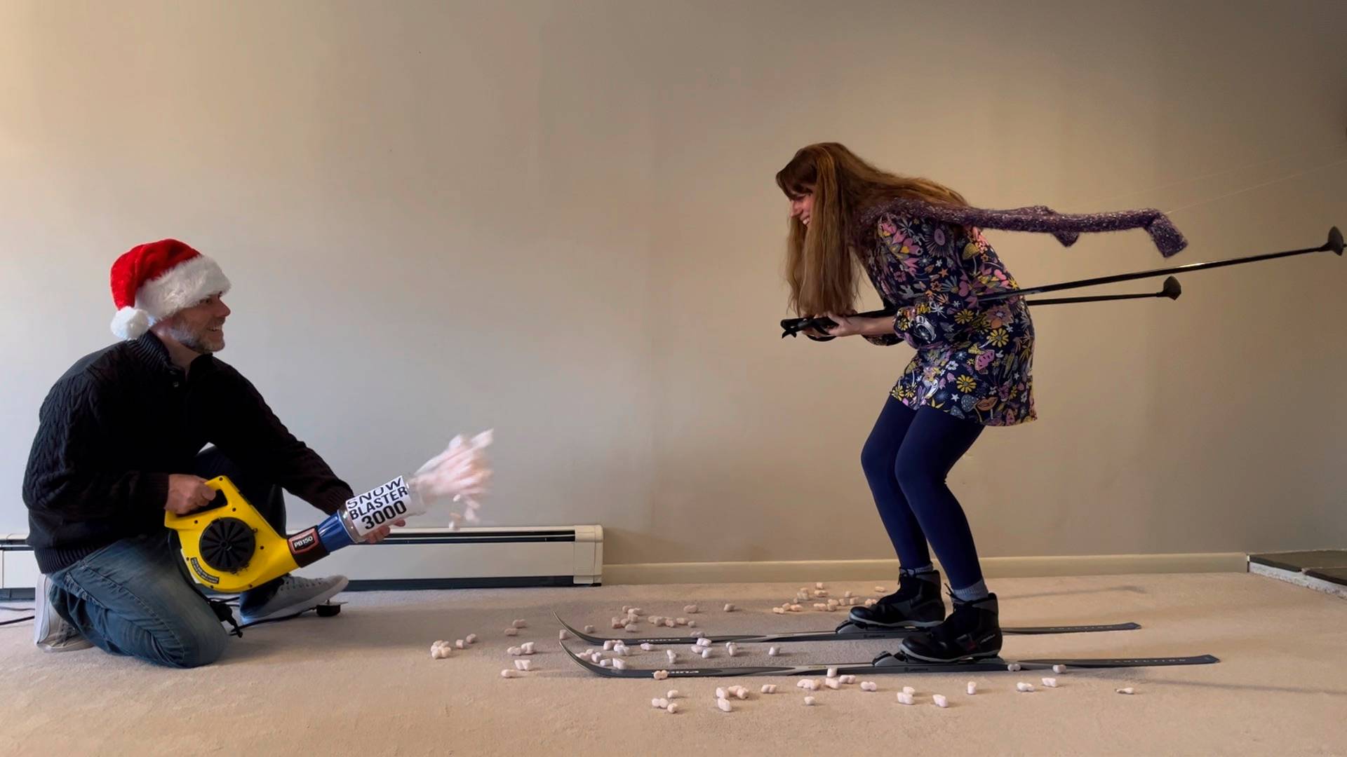 Photo of a man in a Santa hat holding a leaf blower that is blowing styrofoam peanuts at a woman on skis in a living room.