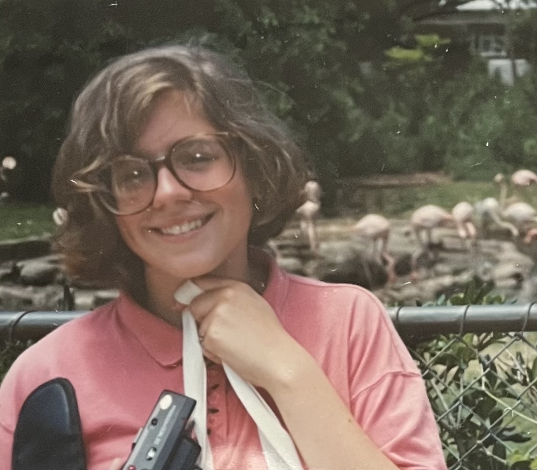Vintage photo of the author wearing very large brown rimmed glasses and a coral pink polo shirt with a fence and flamingos behind her.