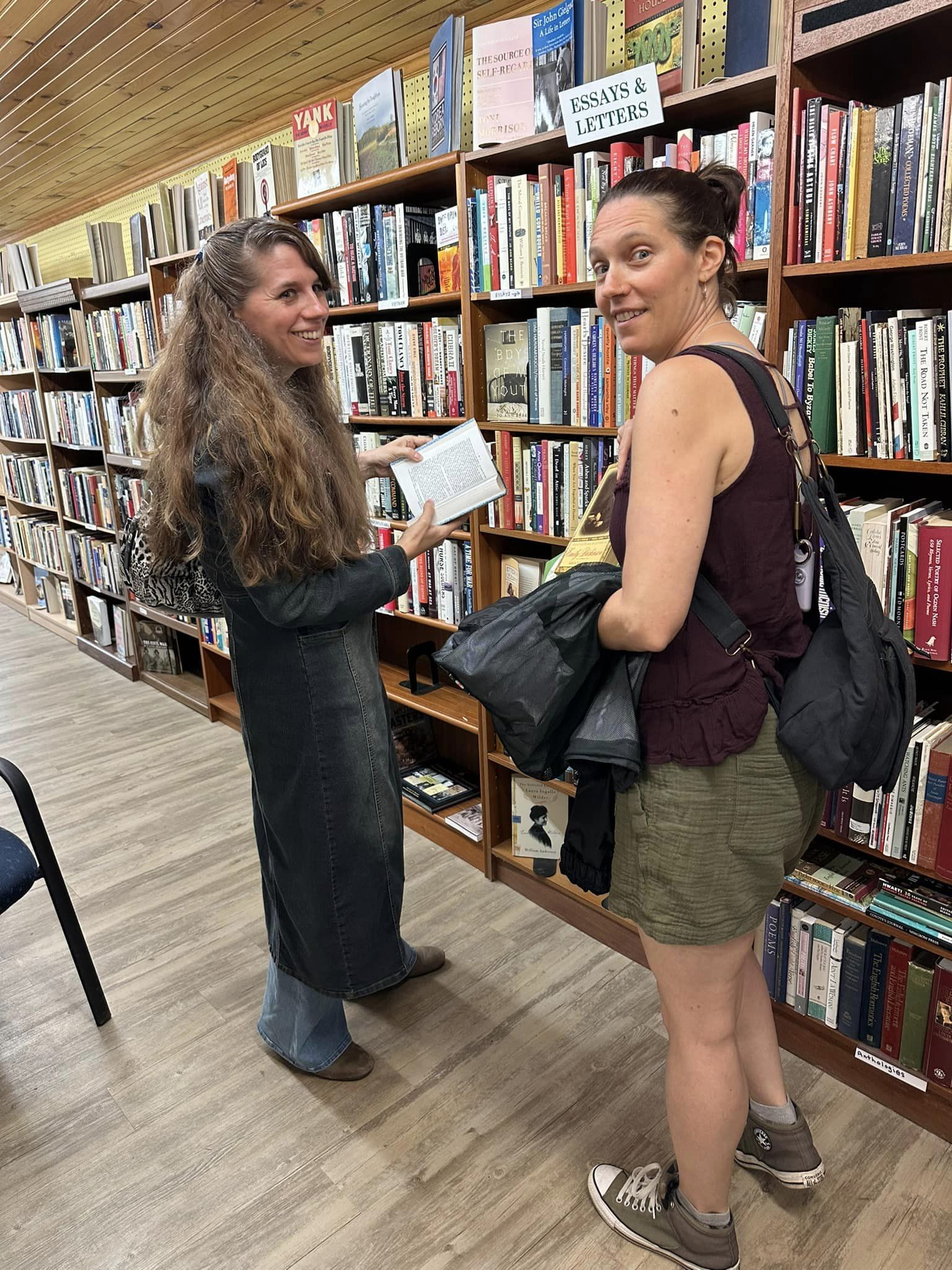 Two women in a bookstore one wearing long jacket and pants and the other wearing a tank top and shorts.