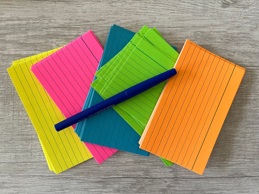 Photo of neon colored index cards and a blue pen on a wood desk.