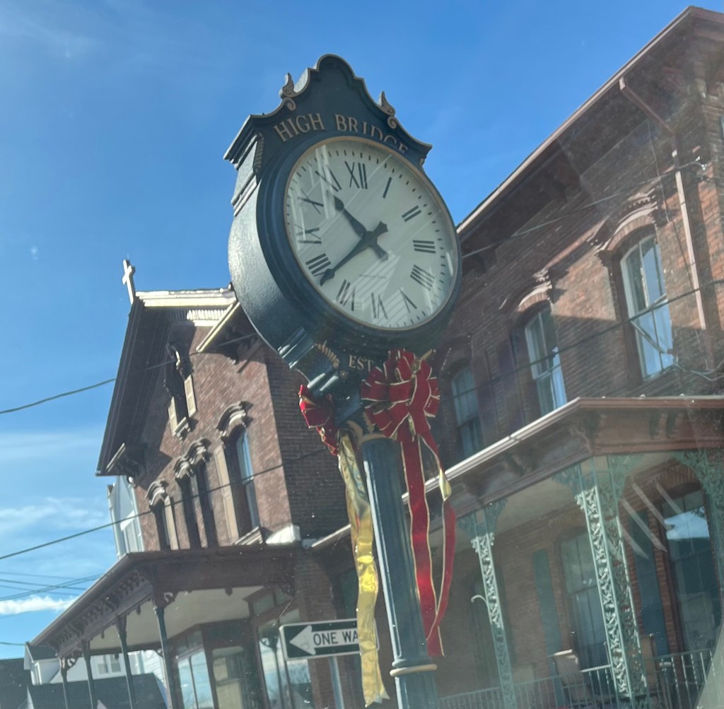 Photo of an old sidewalk clock with a red Christmas bow tied around it, with old brick buildings in the background.