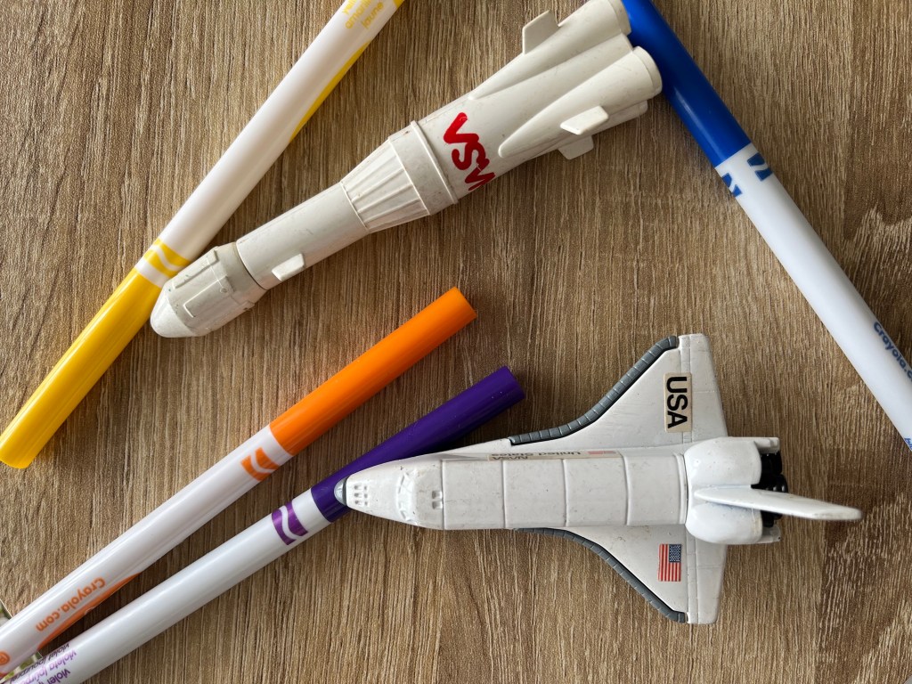 Photo of space shuttle toys and Crayola markers on a wooden surface.