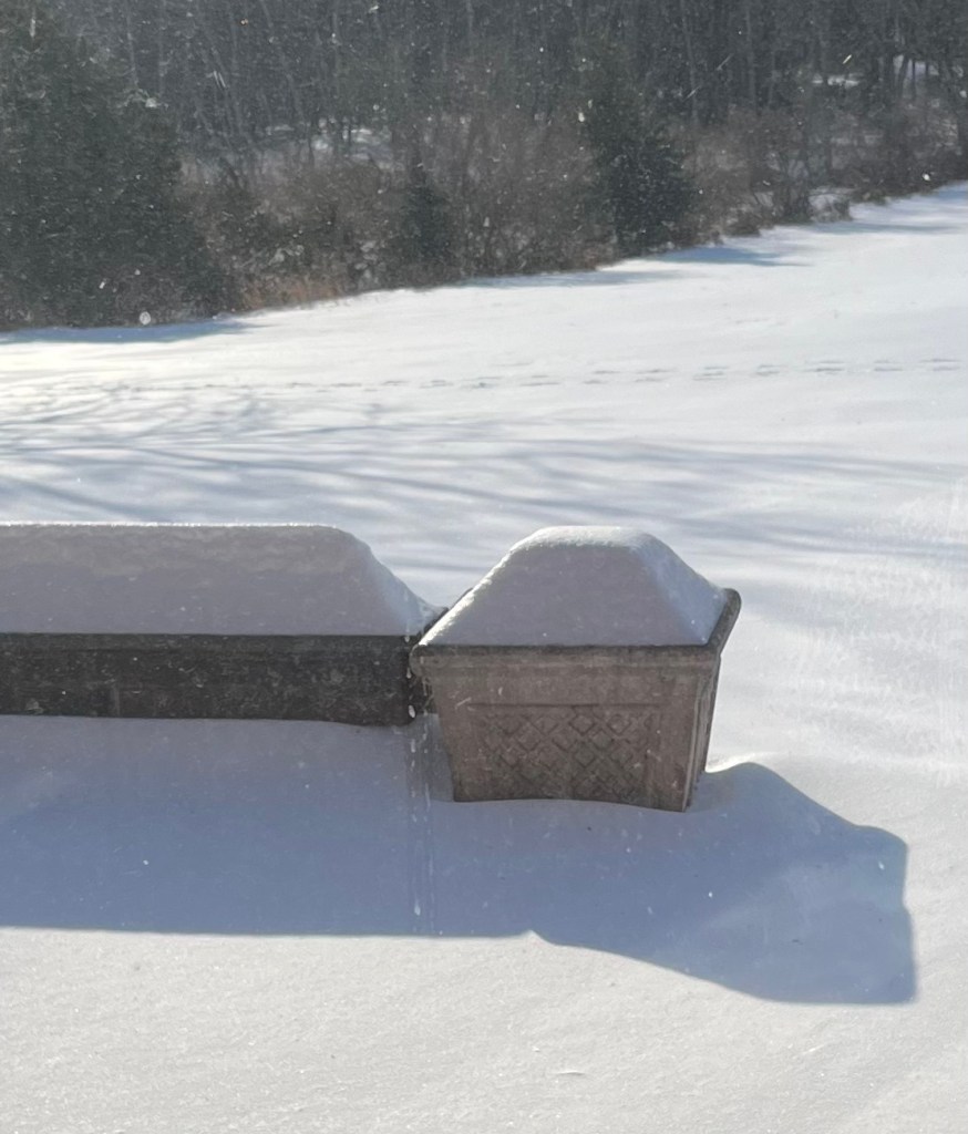 Photo of a snow covered yard including a snow covered patio wall and snow covered planter taken through a very dirty window.