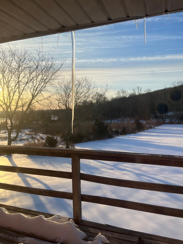 Photo of icicles hanging off a roof with a snowy yard in the background and the rising sun shining.