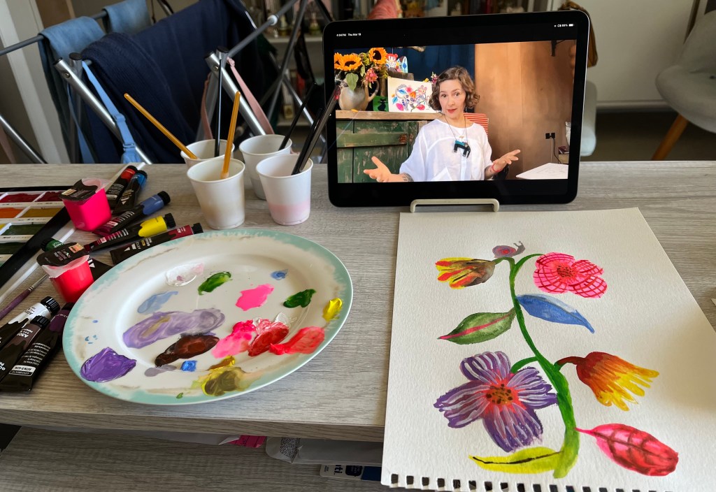 Photo of a desk with paints, a painting palette, a gouache and watercolor floral painting and an ipod showing a video of artist Helen Dardik.
