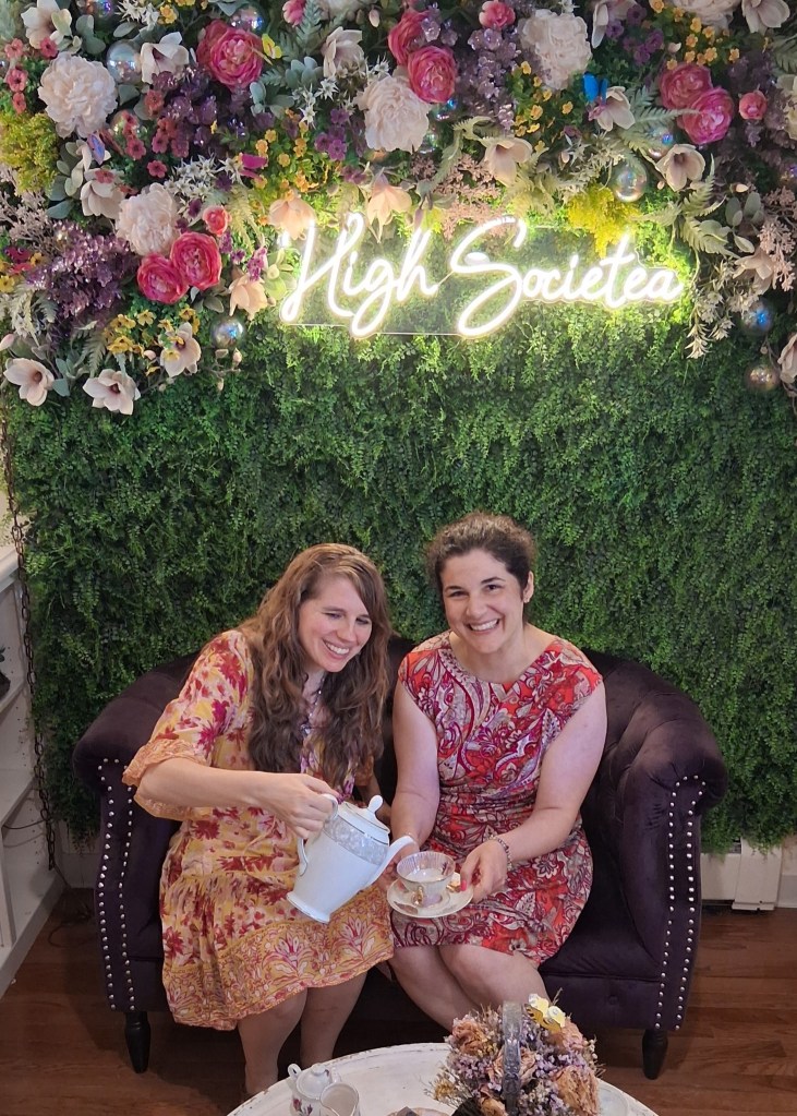 Photo of me and my friend. I am pouring some make believe tea into her cup. Behind us is a botanical decorated wall with a neon sign that reads "High Societea" in script letters.