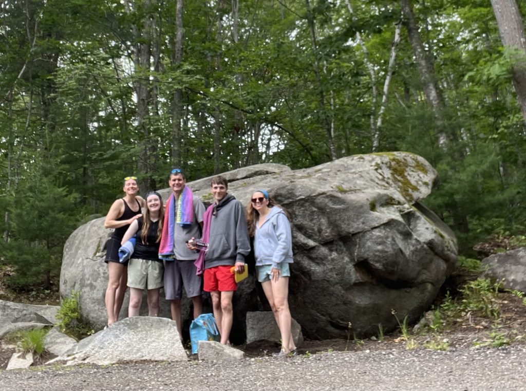 Photo of five people dressed for going swimming standing in front of a large boulder in a wooded area.