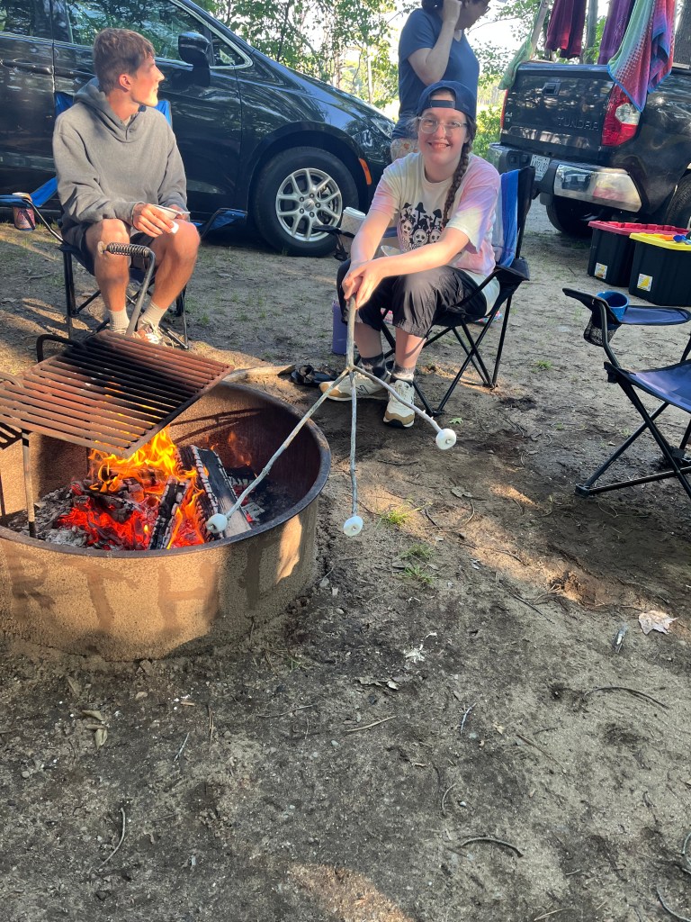 Photo of a young man and a young woman sitting in camp chairs by a camp fire. The young woman is holding a branch with three prongs. There's a marshmallow skewered on each prong.