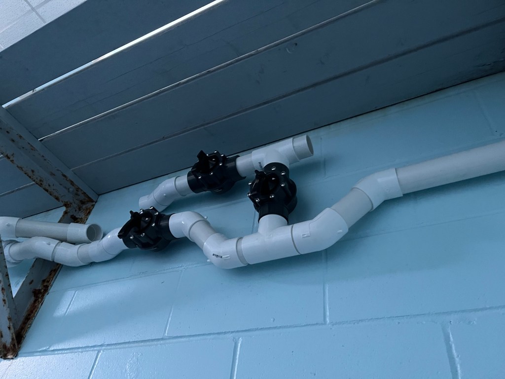 Photo of white PVC pipes and assorted valves mounted on a light blue cinderblock wall.